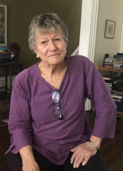 A portrait of Molly Thom, a woman in her 80s, wearing a purple shirt with glasses hanging from her collar.