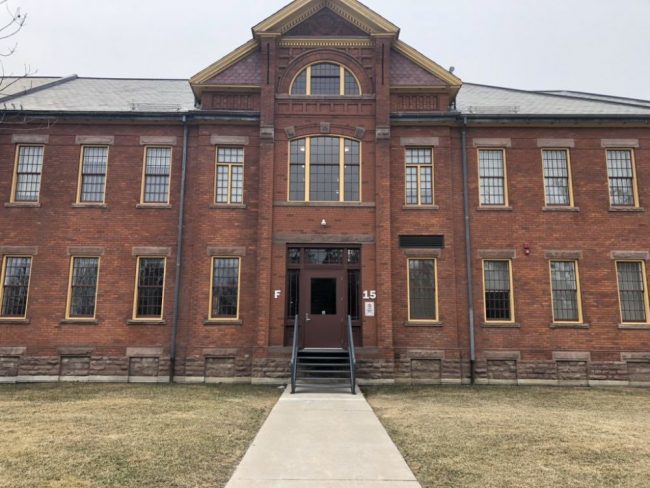 The front of a red brick building with yellow window sills and 'F' on the left side of the door and '15' on the right side in bold grey lettering.