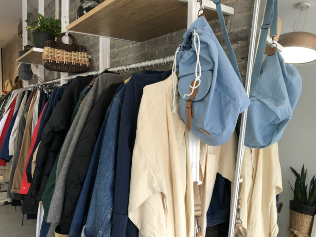 White clothing rack with baby blue bag hanging at the end, an array of neutral colored clothes hanging on the rack.
