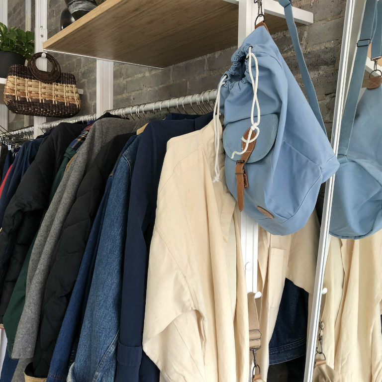 White clothing rack with baby blue bag hanging at the end, an array of neutral colored clothes hanging on the rack.