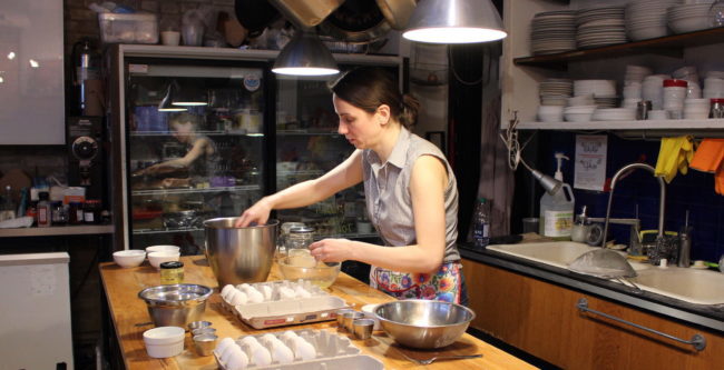 A woman is mixing something in a silver mixing bowl. There are other bows as well as eggs on the table in front of her.