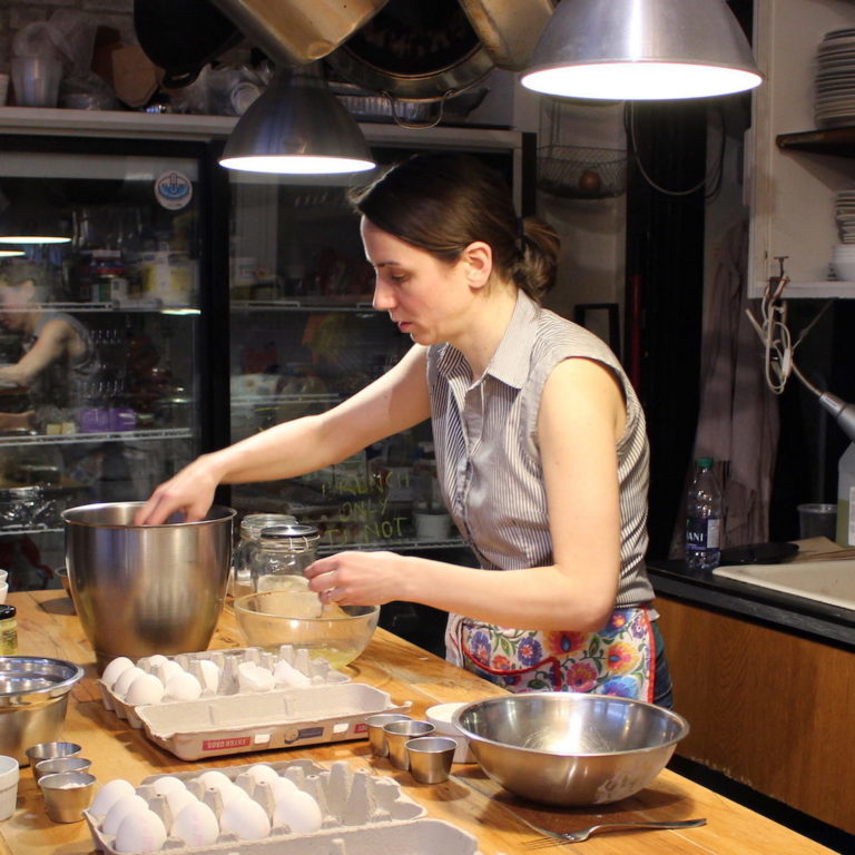 A woman is mixing something in a silver mixing bowl. There are other bows as well as eggs on the table in front of her.