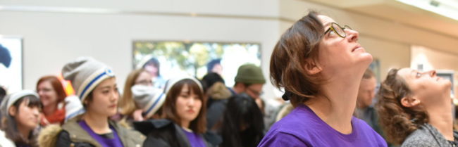 A large group of people standing in a tall room. There is one person in focus, and they are staring upwards at something out of shot.