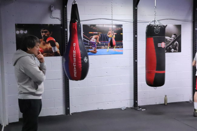a man is standing in front of two punching bags. Behind him there are large wrestling images.