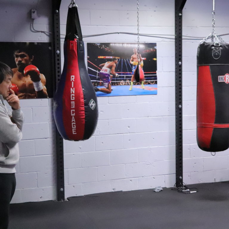 IMG_0852 a man is standing in front of two punching bags. Behind him there are large wrestling images.