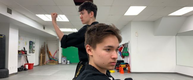 Two people are posed in a fighting stance in a white studio There are workout accessories sitting against a green wall in the background.