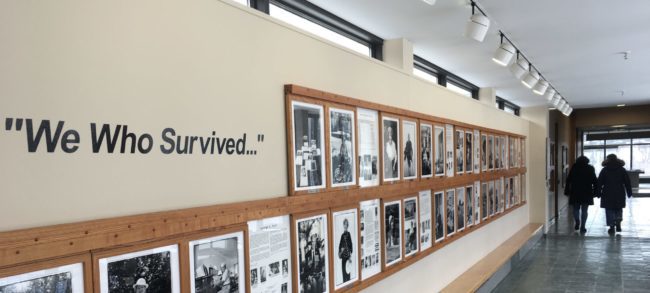 Hallway wall of framed photos of black and white Holocaust survivors. Beside the photos there is a wall vinyl that says "We Who Survived..."