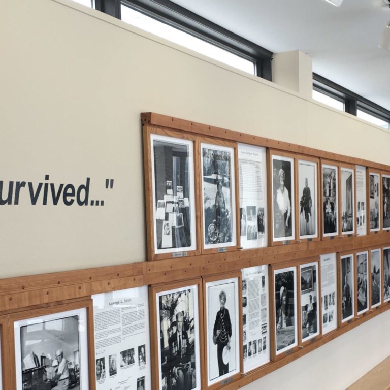 Cover Image; The Sarah and Chaim Neuberger Education Centre’s wall of remeberance Hallway wall of framed photos of black and white Holocaust survivors. Beside the photos there is a wall vinyl that says "We Who Survived..."
