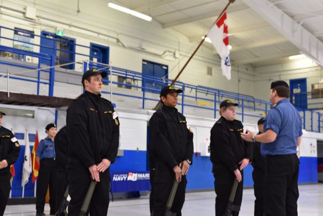 Group of people in uniform doing rifle drill while their instructor is teaching.