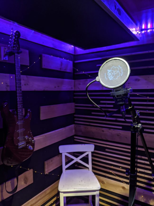 Inside a vocal booth with neon lighting, a mic, a electric guitar and a white chair.