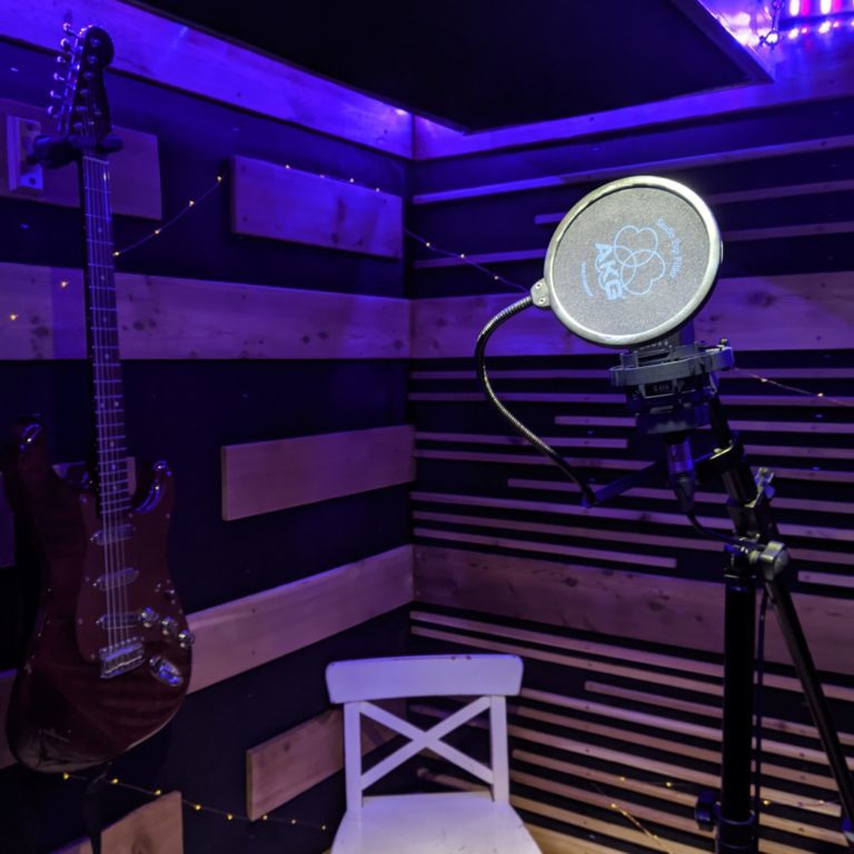 Inside a vocal booth with neon lighting, a mic, a electric guitar and a white chair.