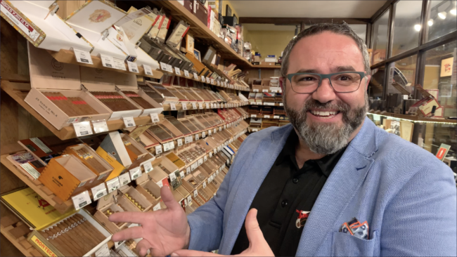 Person with a blue blazer smiling standing in front of shelves of cigars.