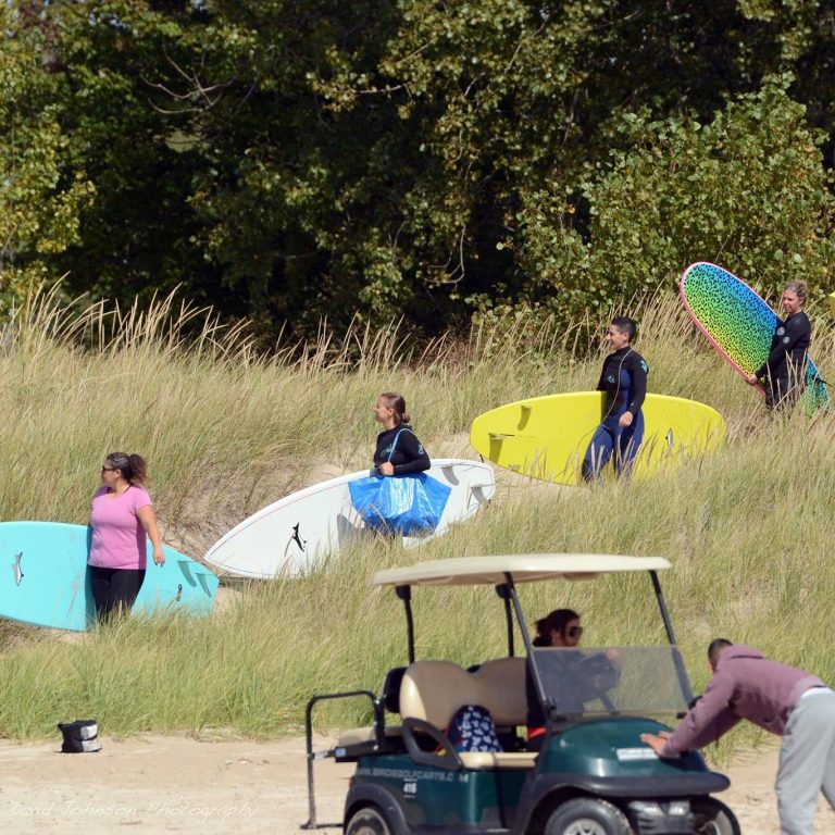 Five people holding surfboards walking down towards a beach.
