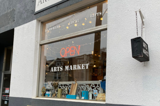 The exterior of the Arts Market. The building is white stucco, and there is a neon open sign hanging in the main window.