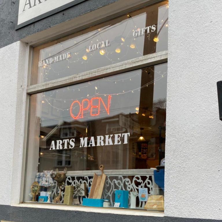 The exterior of the Arts Market. The building is white stucco, and there is a neon open sign hanging in the main window.