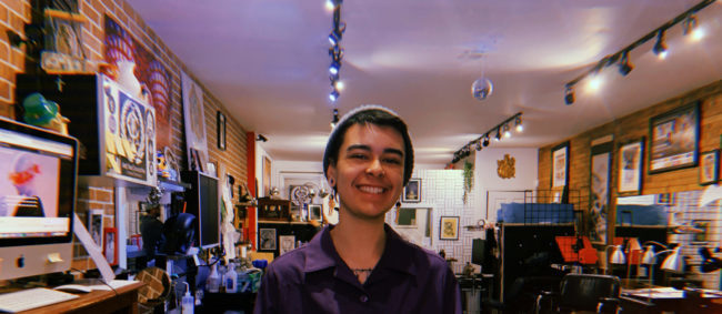 a person smiling at the camera, behind him a brick-walled room is brightly lit. The room is busy but filled with life.