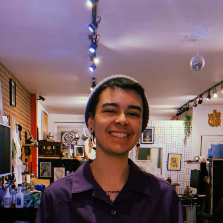 a person smiling at the camera, behind him a brick-walled room is brightly lit. The room is busy but filled with life.