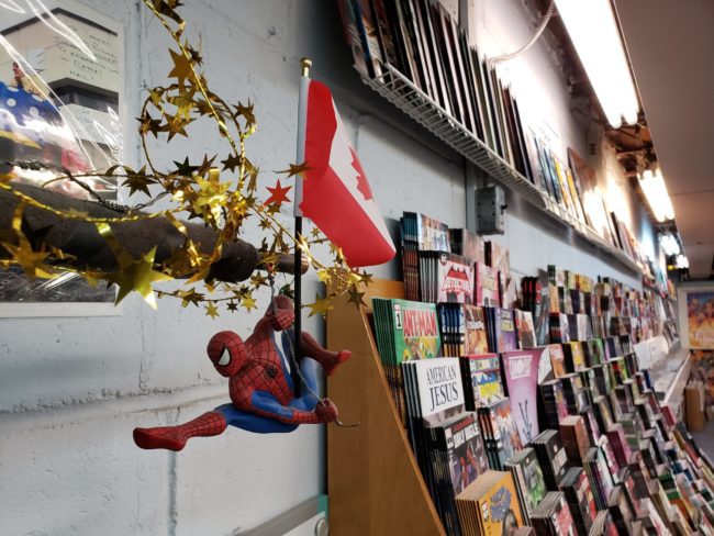 Spiderman hanging off a small canadian flag. in the background there are large sloping shelves filled with comic books.