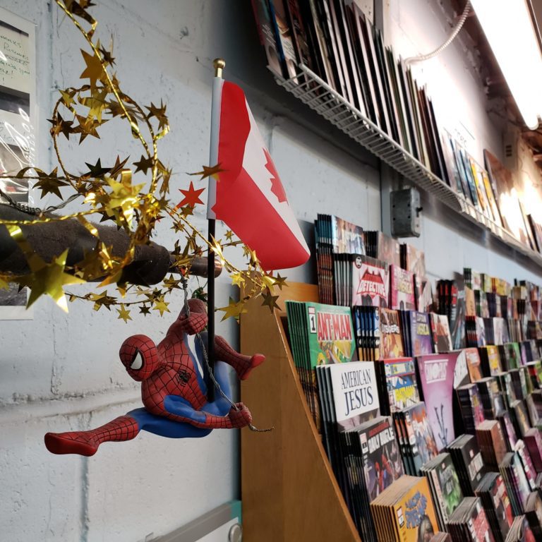 20200227_154241 Spiderman hanging off a small canadian flag. in the background there are large sloping shelves filled with comic books.