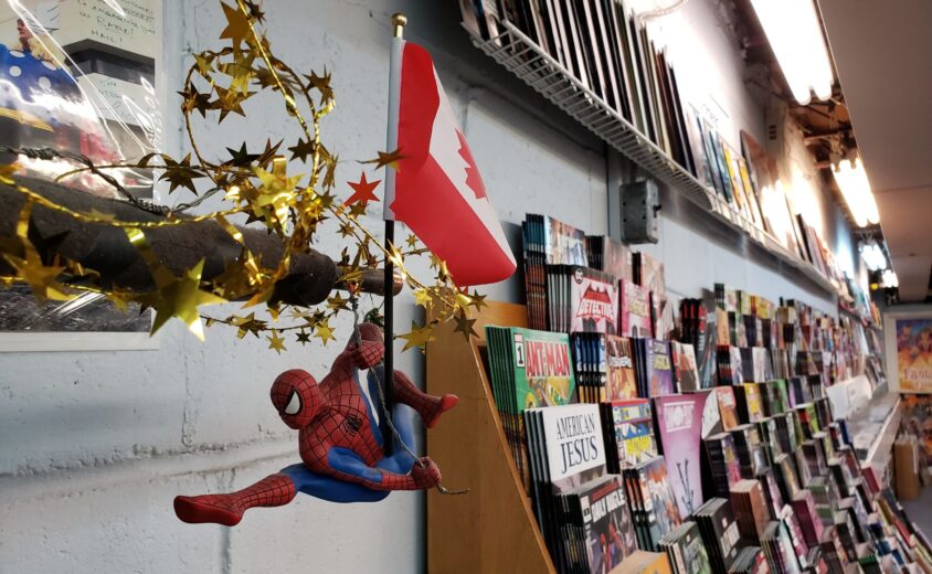 Spiderman hanging off a small canadian flag. in the background there are large sloping shelves filled with comic books.
