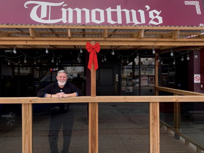 Person standing and smiling under a wooden frame. The frame is laced with hanging Edison bulbs. Above there is a red sign that reads Timothy's.
