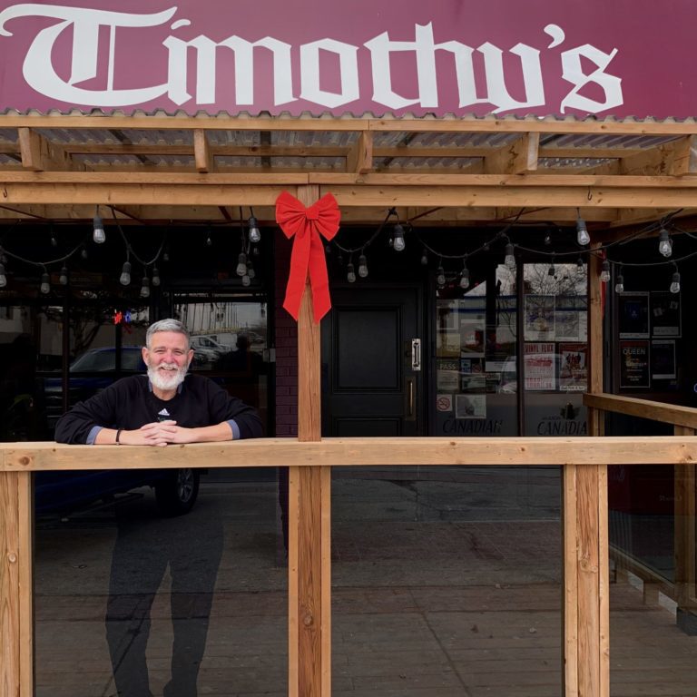 Person standing and smiling under a wooden frame. The frame is laced with hanging Edison bulbs. Above there is a red sign that reads Timothy's.