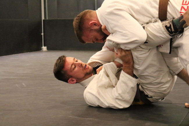 Two people fighting in white gi's during a competition. One person is on his back with legs in a defensive position. The other person is leaning against the other person's legs.