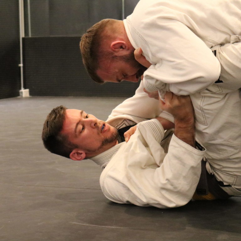 Academy-One-1 Two people fighting in white gi's during a competition. One person is on his back with legs in a defensive position. The other person is leaning against the other person's legs.
