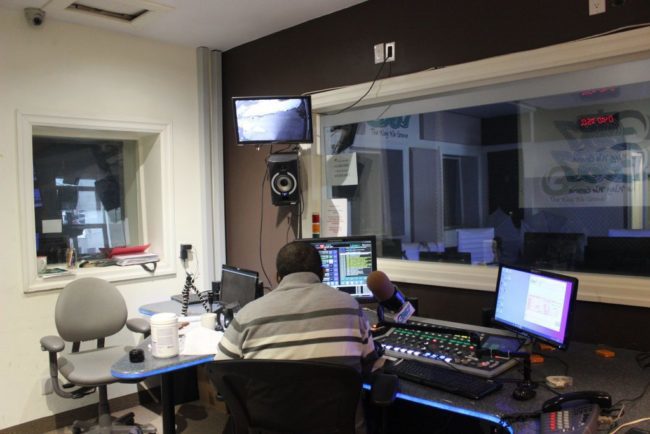 a wide shot of a radio setup. There is someone sitting with their back to the camera, preparing to talk into a large microphone beside them.