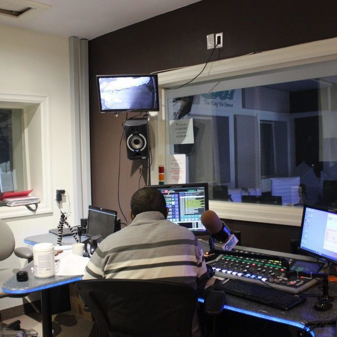 a wide shot of a radio setup. There is someone sitting with their back to the camera, preparing to talk into a large microphone beside them.