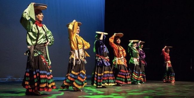 Women dancing onstage in silk shirts and brightly coloured skirts. They're holding wicker baskets on top of their heads.