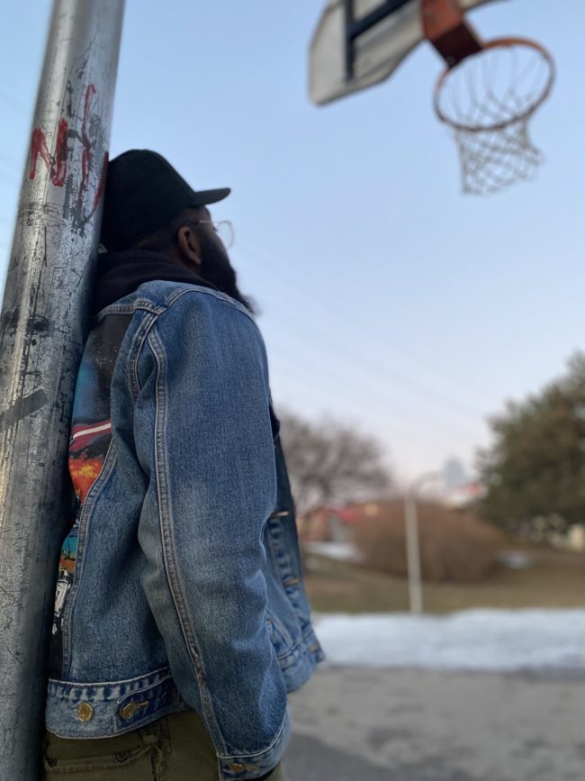 Person in a black hat and a jean jacket leaning against a basketball stand.