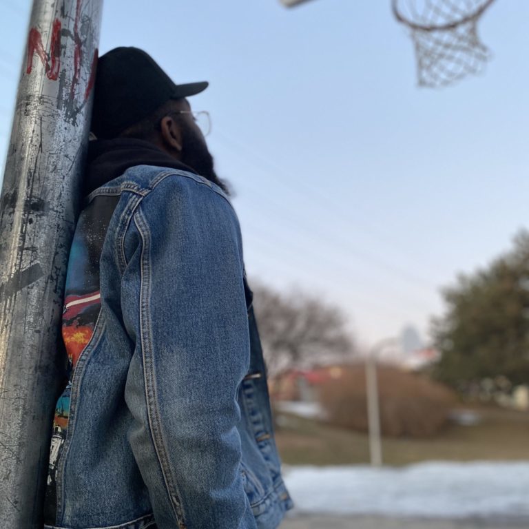 IMG_0761 Person in a black hat and a jean jacket leaning against a basketball stand.