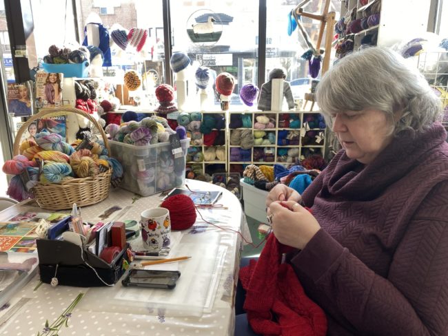 A woman is sitting at a table, knitting with some red yarn. The table and the surrounding area are completely covered in yarn balls, and the table itself is covered in a polka dot cloth.
