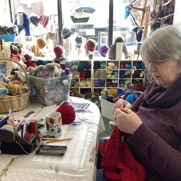 A woman is sitting at a table, knitting with some red yarn. The table and the surrounding area are completely covered in yarn balls, and the table itself is covered in a polka dot cloth.