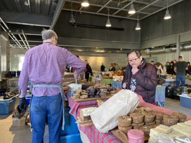 Farmers market. There are people throughout the market looking at baked goods and other various food products.