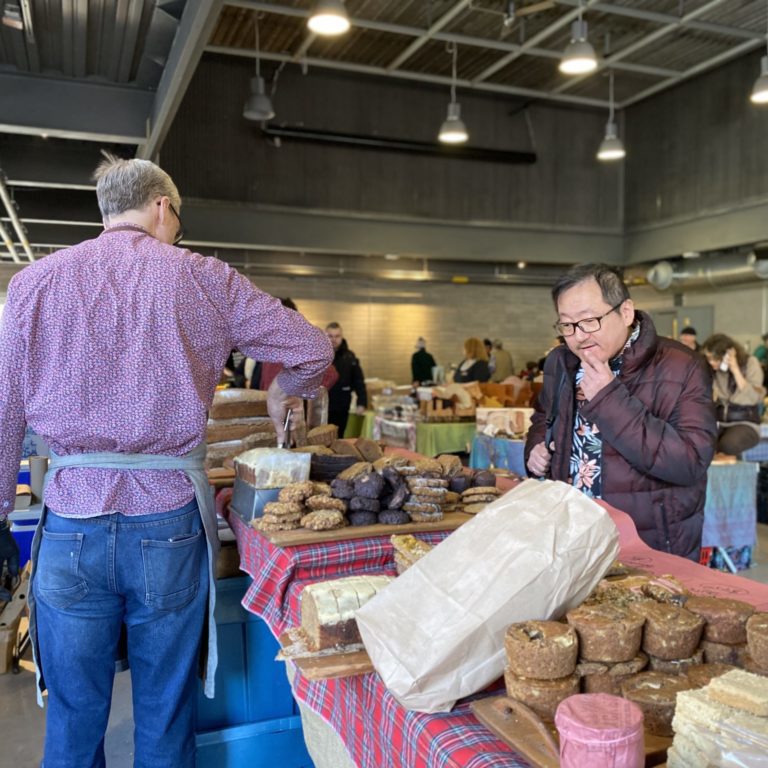 Farmers market. There are people throughout the market looking at baked goods and other various food products.