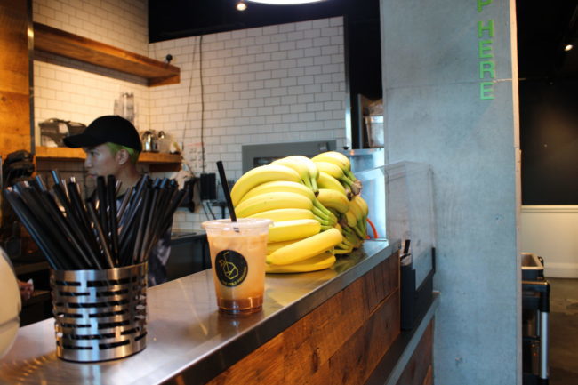 Iced drink sitting on a stainless steel countertop. A bunch of bananas sits in the background next to some black straws in a metal container.