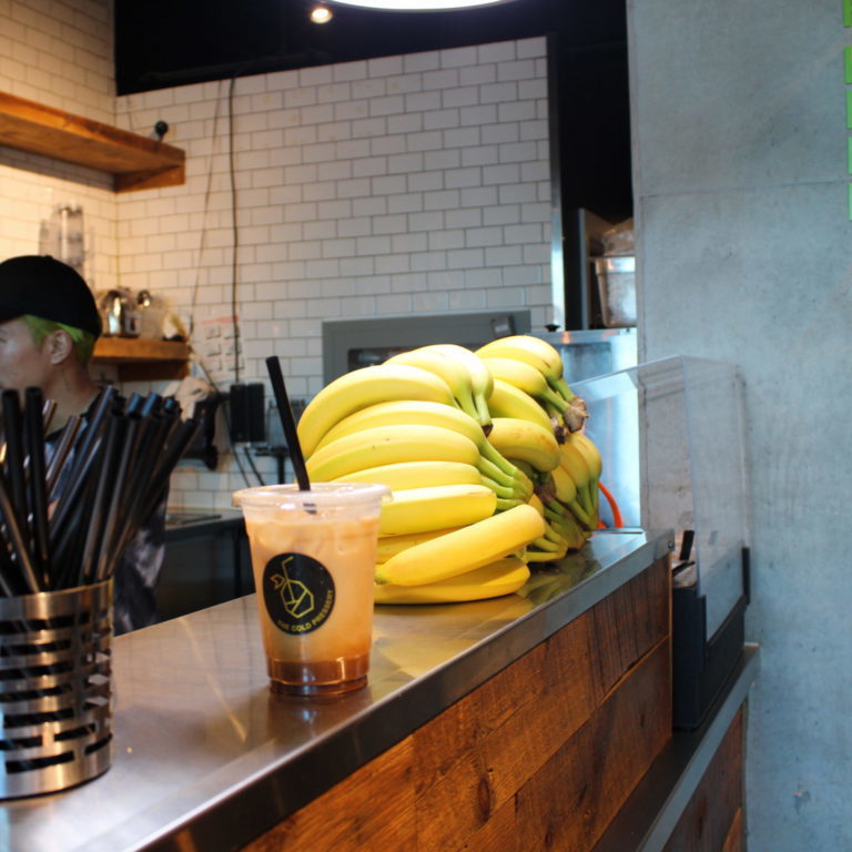 Iced drink sitting on a stainless steel countertop. A bunch of bananas sits in the background next to some black straws in a metal container.