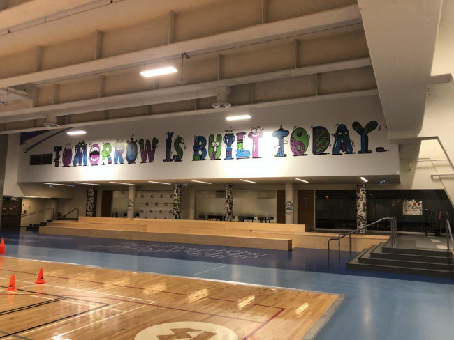 Gym floor and bleachers. The sign above the bleachers says tomorrow is built today in blue, green, and purple animated letters.