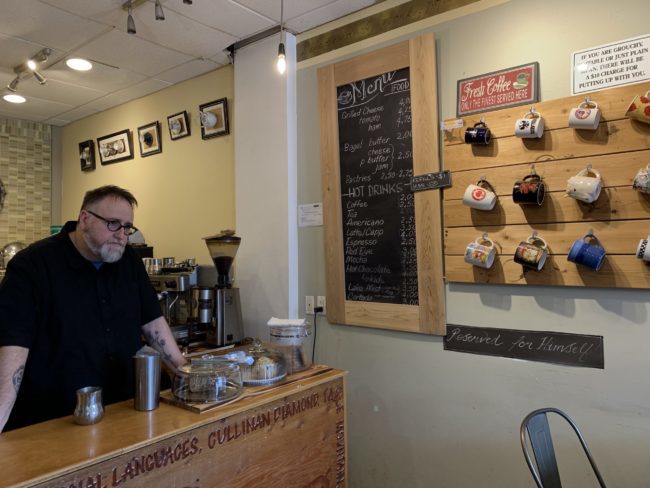 a man stands at the end of a coffee bar, looking pensive. There is a food and drink menu on the wall, beside a hanging mug board. Underneath the mugs there is a blackboard sign that says 'reserved for himself!'