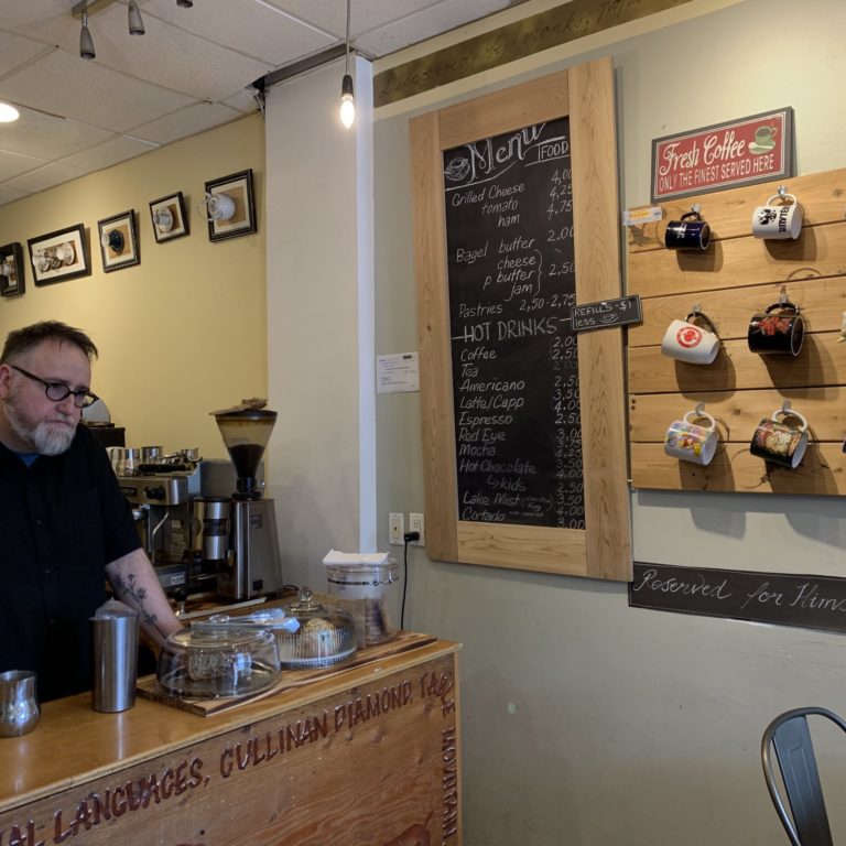 a man stands at the end of a coffee bar, looking pensive. There is a food and drink menu on the wall, beside a hanging mug board. Underneath the mugs there is a blackboard sign that says 'reserved for himself!'