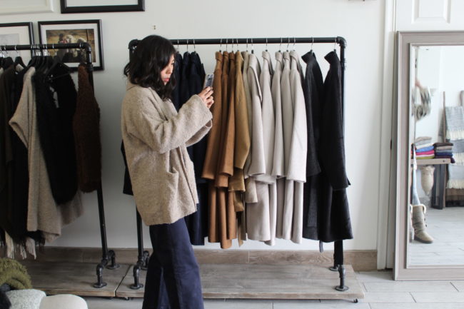 A person is standing and taking a photo with their phone. Behind them there are long coats in blacks, whites, and browns.