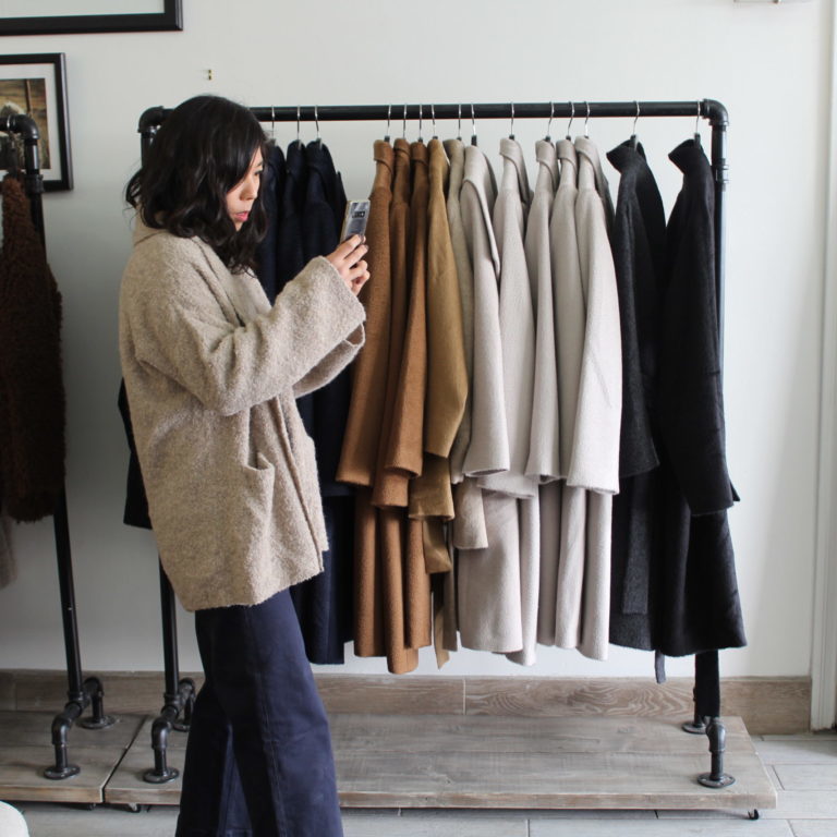 A person is standing and taking a photo with their phone. Behind them there are long coats in blacks, whites, and browns.