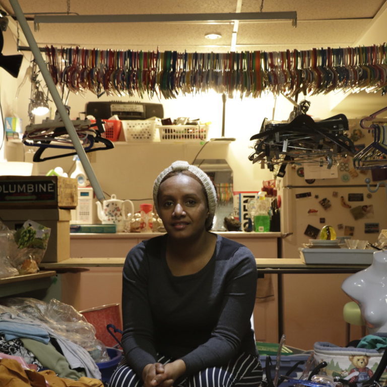 Person sitting next to a pile of clothes. Behind her are thousands of clothes hangers.