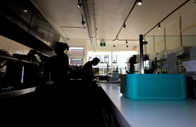 A long shot of two people working behind a food counter. There is light coming into the large front windows, partially obscuring those working in shadow.