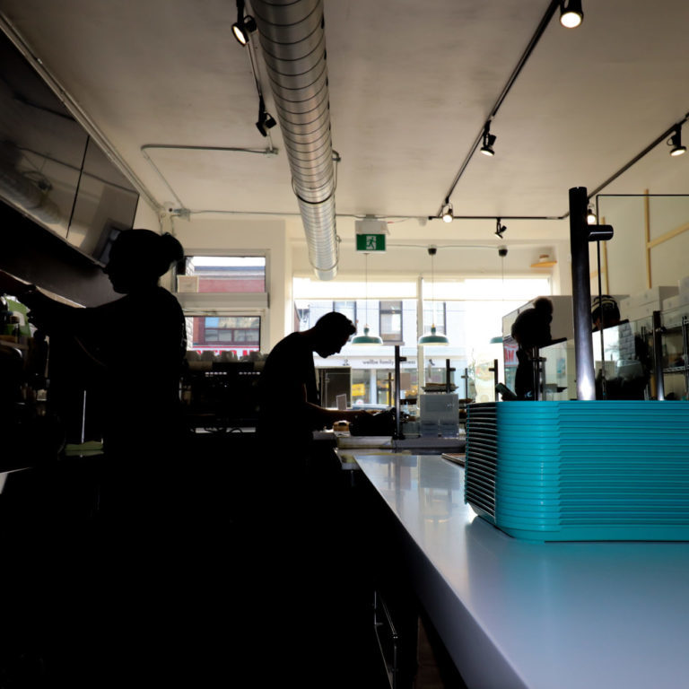 A long shot of two people working behind a food counter. There is light coming into the large front windows, partially obscuring those working in shadow.