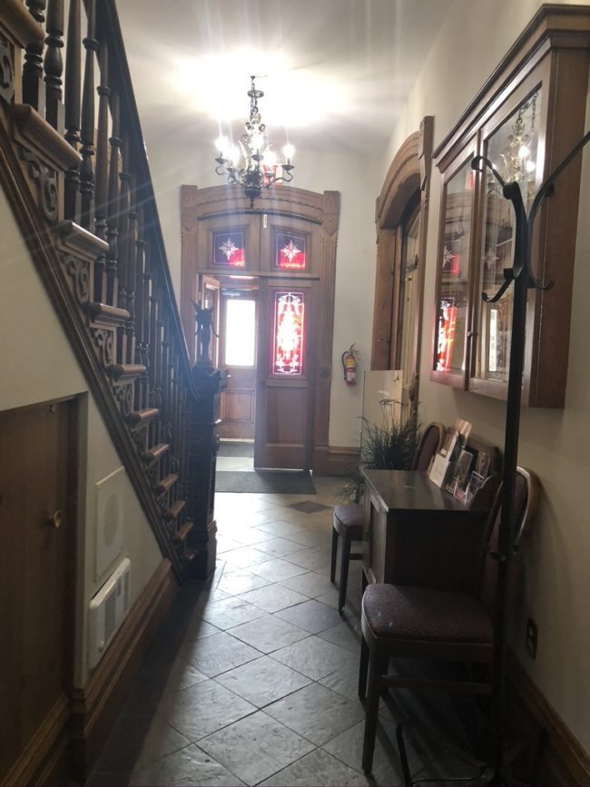 Narrow hallway leading to a front door. The walls are white with a beige tiled floor. To the left there is a wooden staircase and to the right another entranceway.