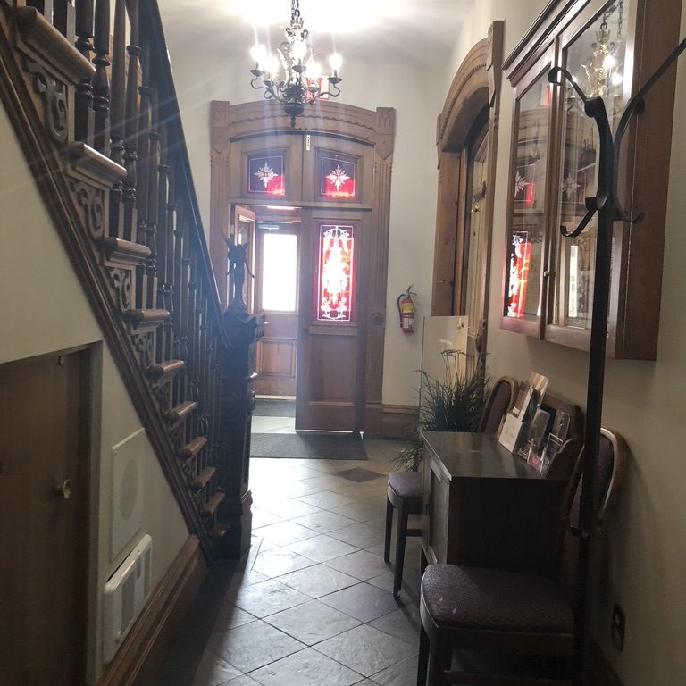 Narrow hallway leading to a front door. The walls are white with a beige tiled floor. To the left there is a wooden staircase and to the right another entranceway.