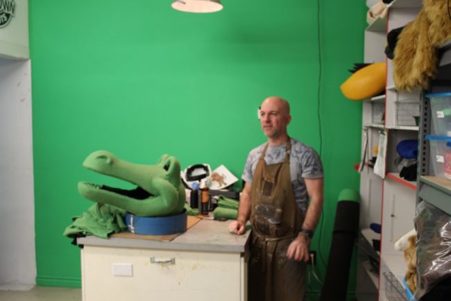 Man in a grey t-shirt and a leather apron next to a mascot crocodile head in a green room.